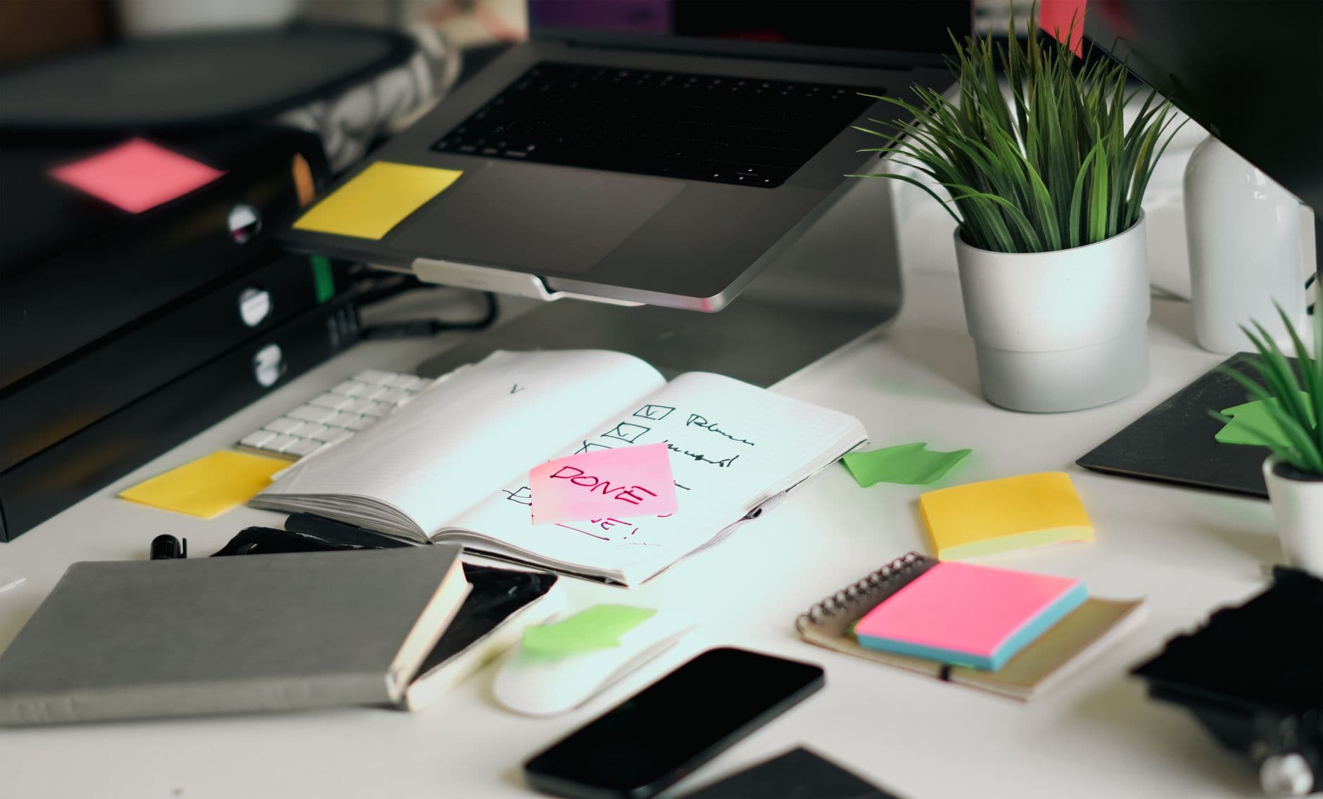 Laptop, notebooks, checklists, and colorful sticky notes across a busy office desk.